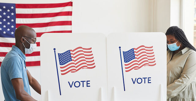 图为两名戴着口罩在投票站投票的人，他们身后的投票站上悬挂着美国国旗